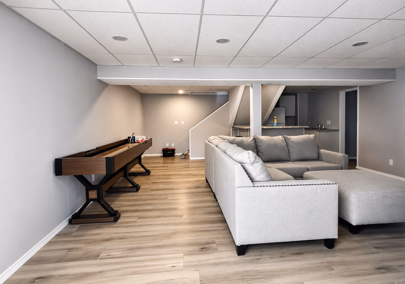 Modern basement with light wood floor, gray L-shaped sofa, shuffleboard table, and kitchenette under stairs.