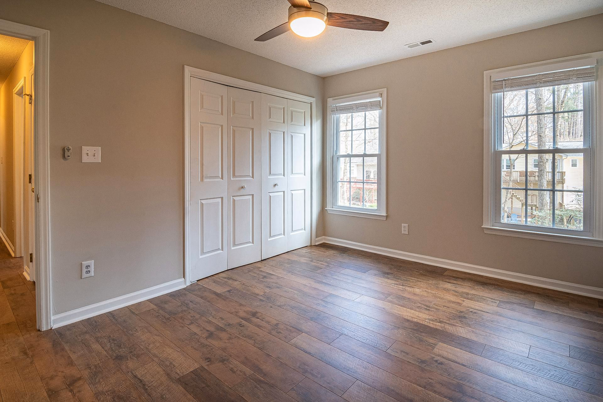 Empty room with wood flooring, two large windows, white closet doors, and a ceiling fan with light.